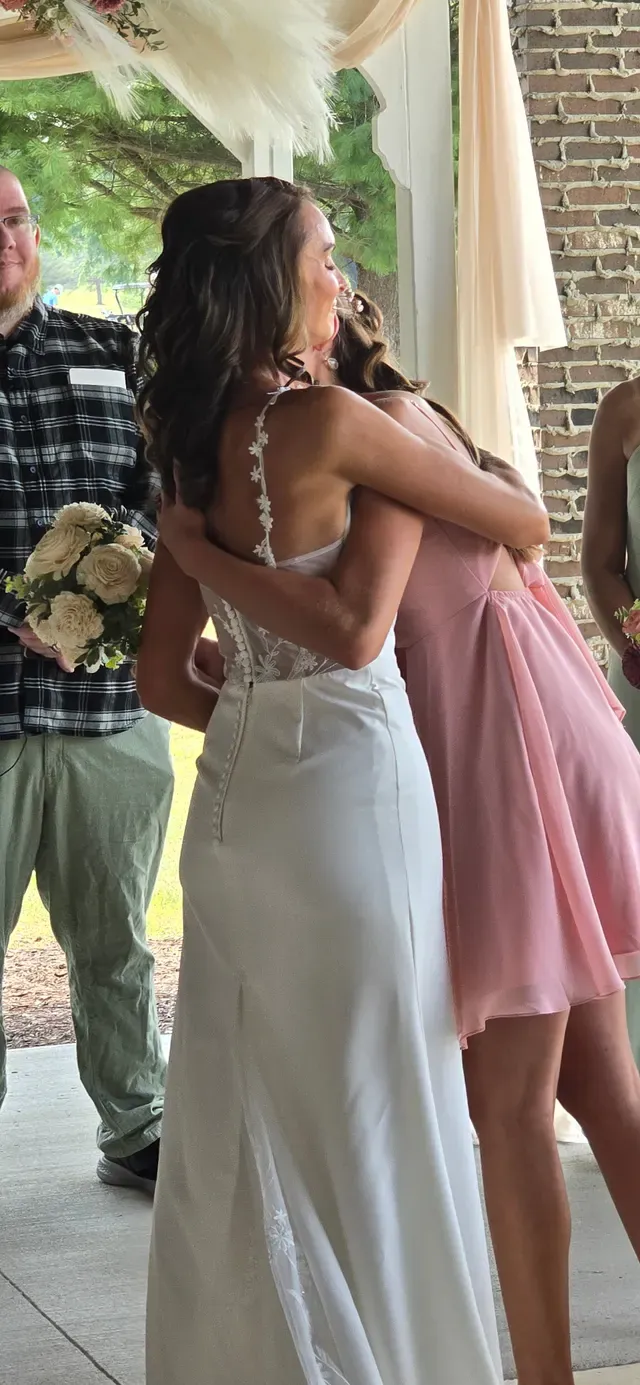 A bride holding a bouquet embracing a guest, seen from behind