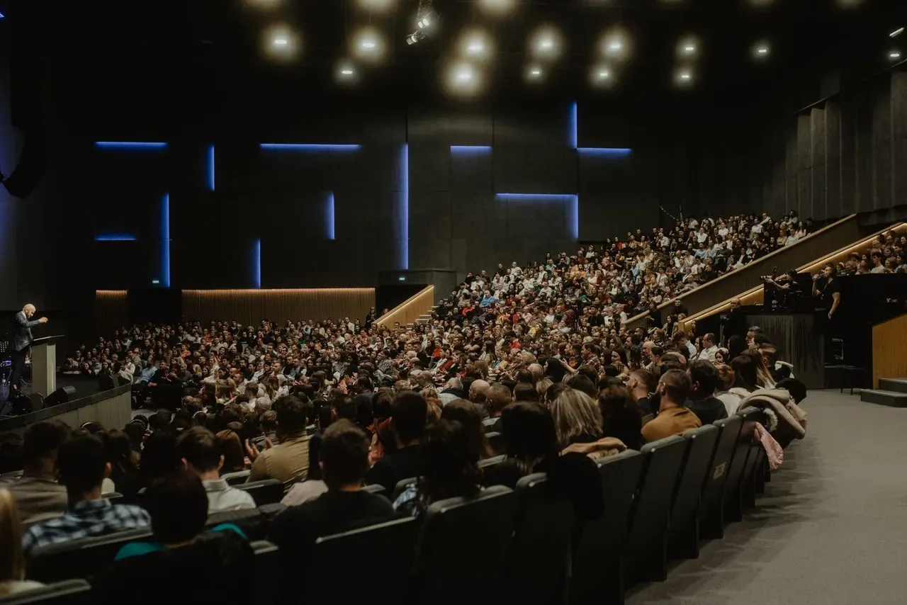 A full auditorium seen from the back, the audience lit blue against a dark wall