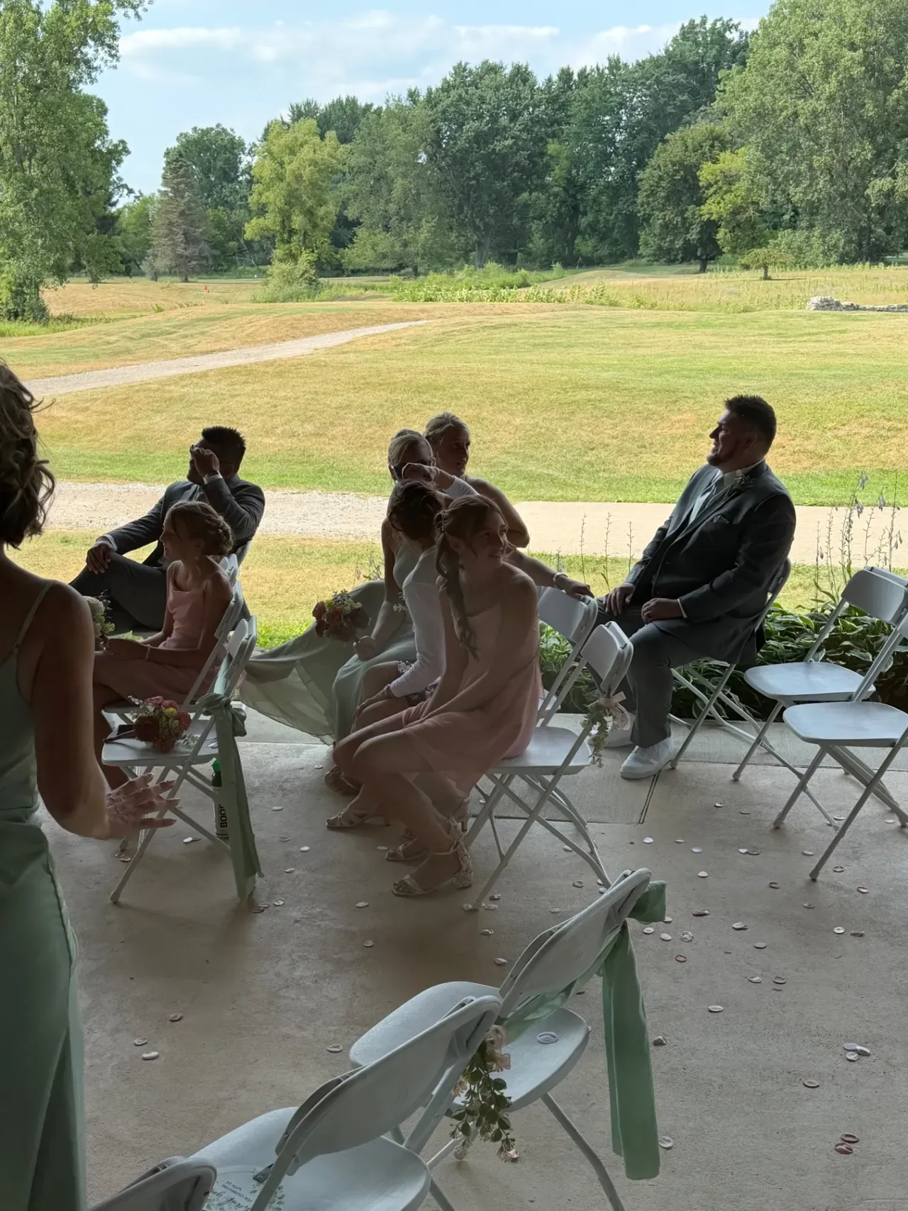 Guests seated on white chairs under a veranda, with a mown fairway and cart path stretching away behind them