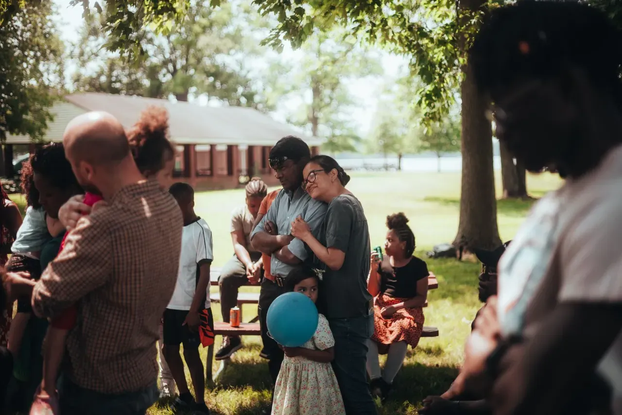 People gathered on a lawn under trees on a bright day, two of them embracing