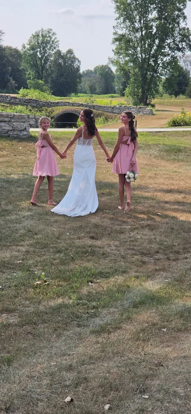 A bride walking across grass holding hands with two flower girls, seen from behind